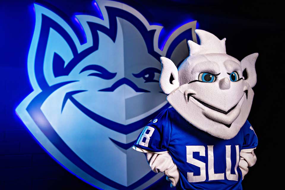 The Billiken mascot poses in front of the Billiken logo.