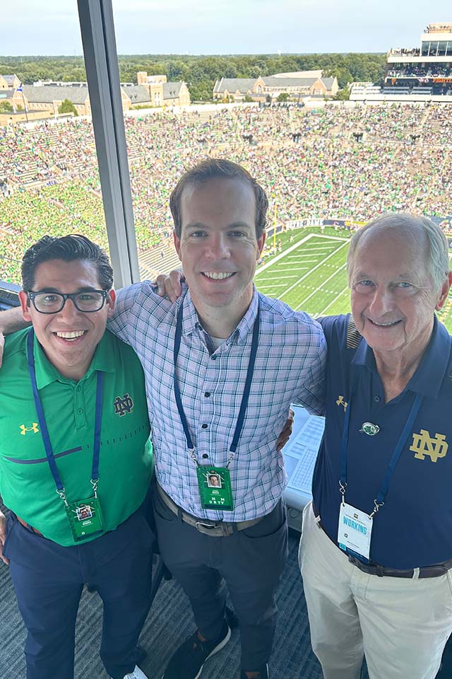 Three men pose for a photo with a stadium behind them. Two of the men are wearing Notre Dame shirts.