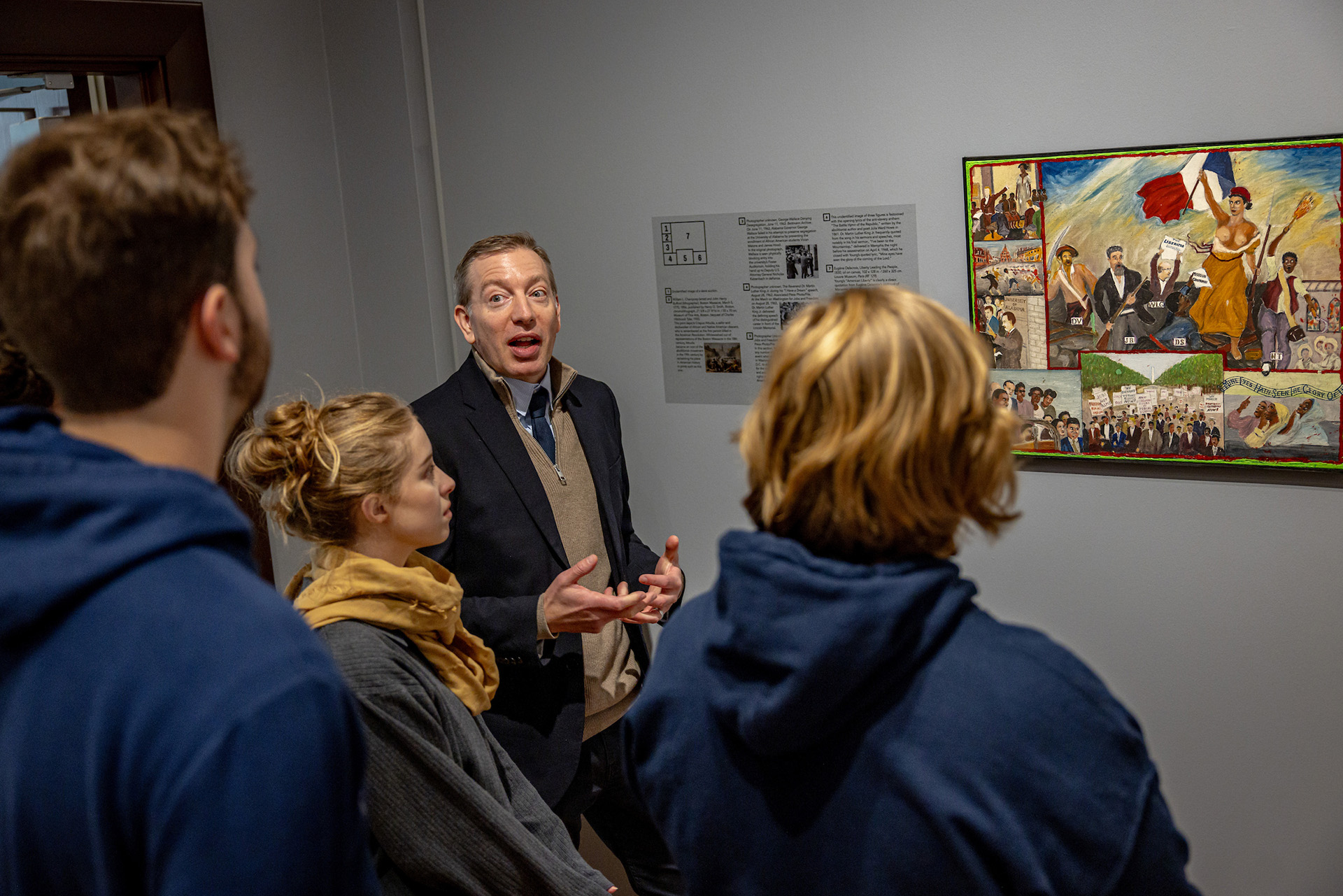 A professor discusses a painting on the wall while students observe.