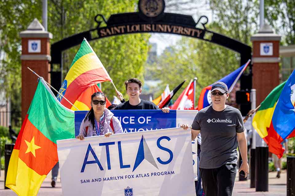 Students holding a sign reading Sam and Marilyn Fox Atlas Program in front of a parade of flags