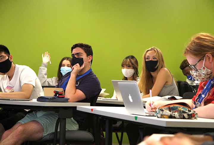 Students sitting at classroom tables in masks look ahead. One student raises her hand.