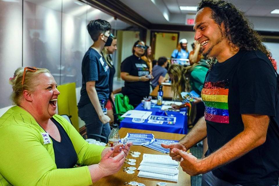 A student wearing a t-shirt with the rainbow flag talks and laughs with another student who is sitting at a table. The table has billiken keychains and other materials.