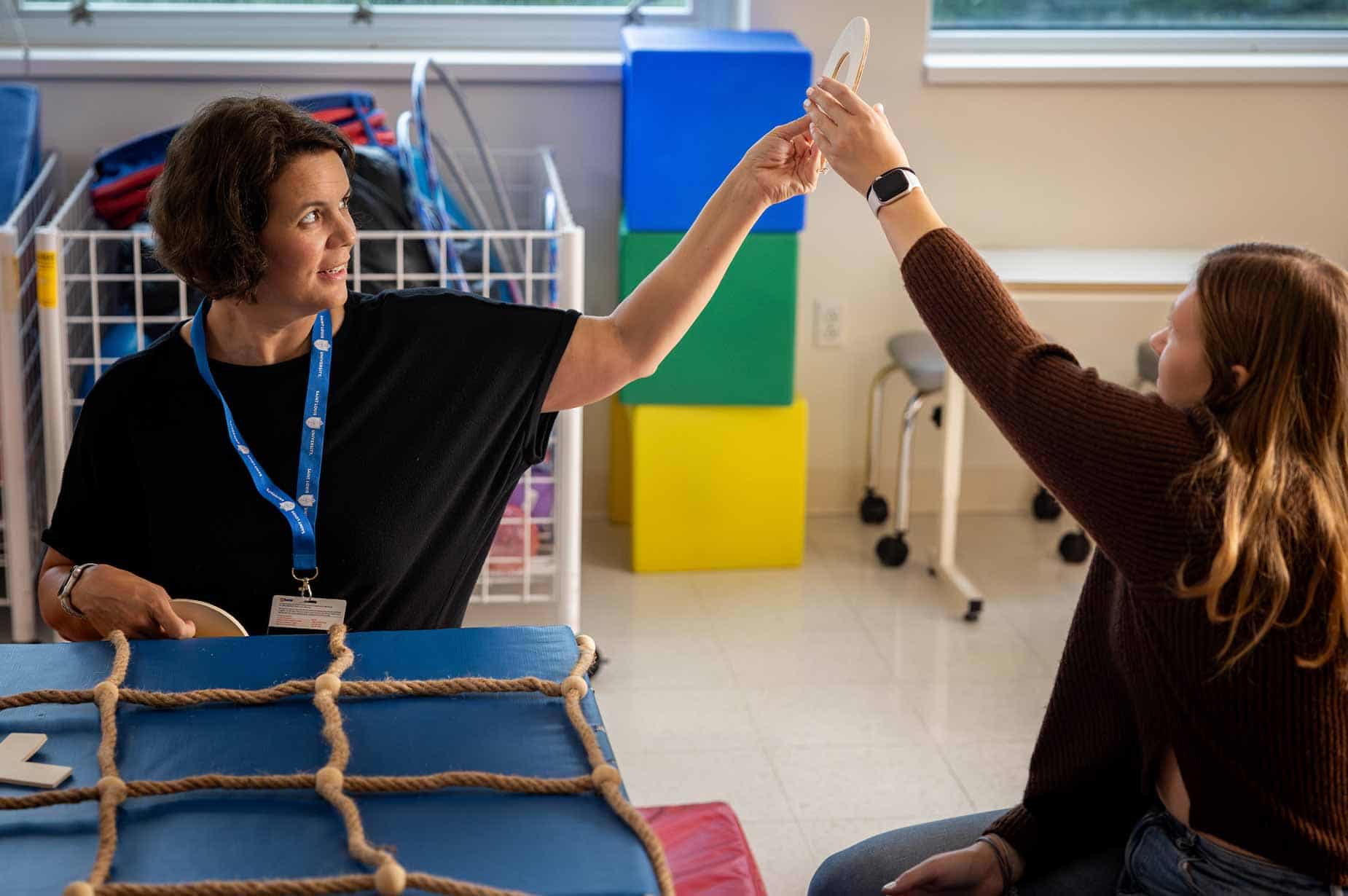 A female professor holds up a wooden O for a student. They’re both sitting in an occupational therapy lab and large colorful mats and blocks can be seen in the background.