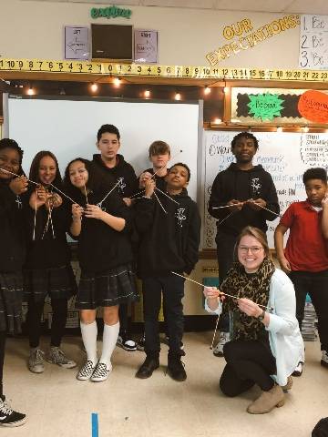 A teacher kneels on the floor of a classroom, with students in the background. They're all holding some sort of sticks in their hands and smiling.