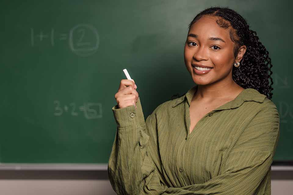 A girl poses for a photo holding chalk in front of chalk board.