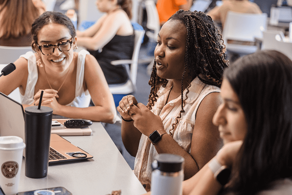 Three female students sit at a table and look at a laptop as they study.