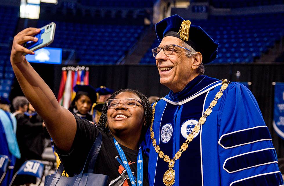 President Fred Pestello wearing a full cap and gown smiles for a selfie with a student