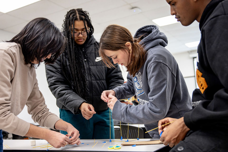 Four engineering students bend over a table working on a project involving wooden sticks.