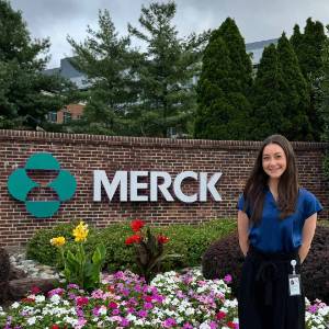 Lauren Favell stands in front of a flower bed and a large sign that says Merck.