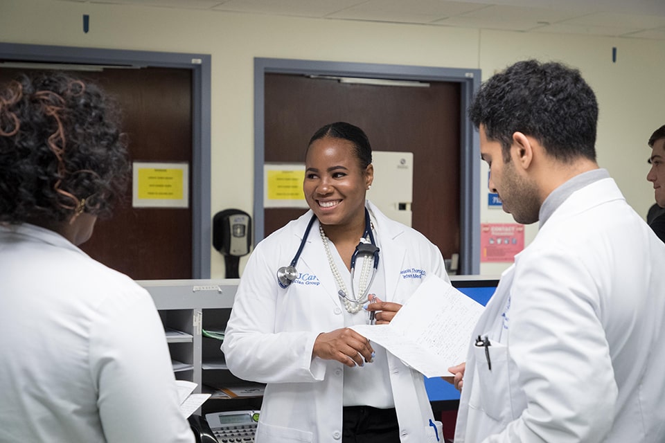 Three students wearing doctors' coats and stethoscopes stand in a hospital and talk.