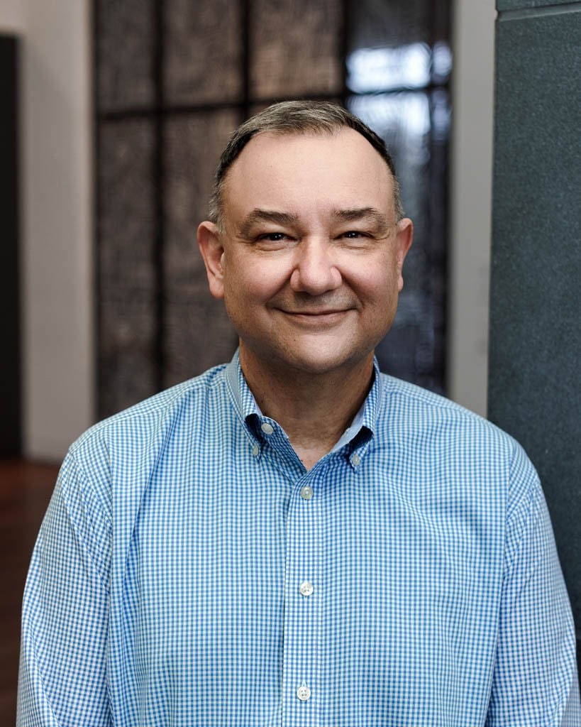 A photograph of curator René Paul Barilleaux, wearing a blue checkered dress shirt and standing in front of a building 