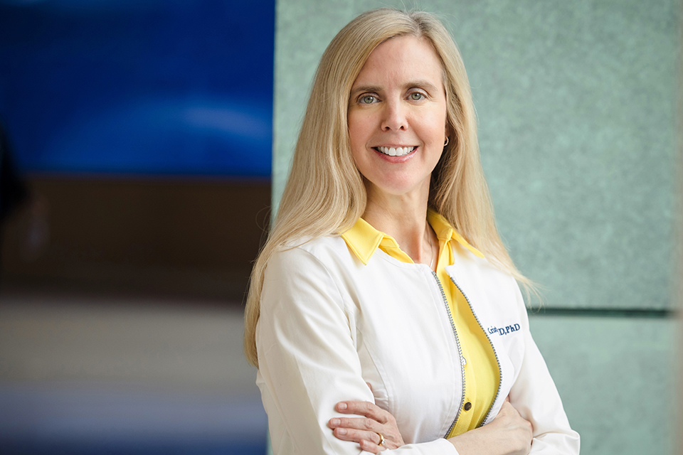 Dr. Lentine poses for a photo. She is wearing a white lab coat and a yellow shirt. Her arms are crossed.