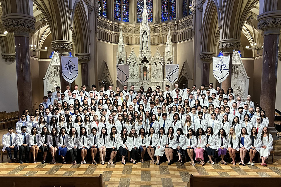 Formally dressed incoming medical students wearing white coats pose for a group photo inside a church.