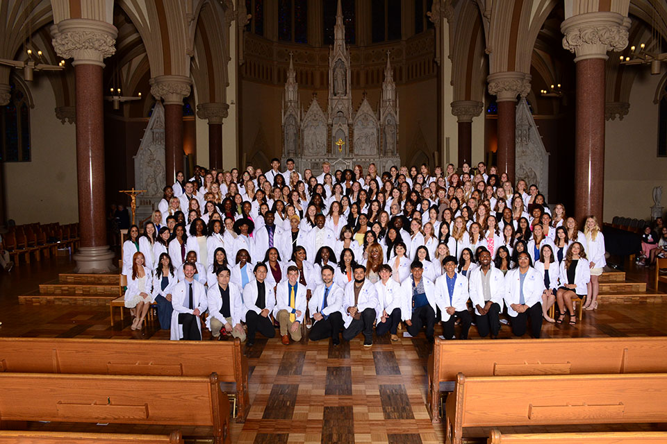 A large group of nursing students wearing white coats pose for a photo inside a dimly lit church.