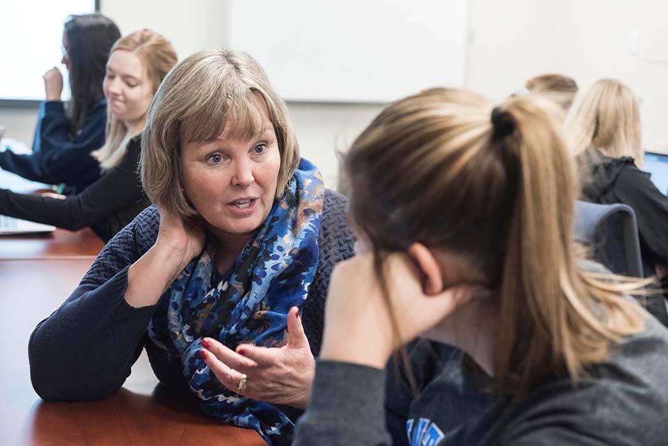 A middle-aged woman with grey hair speaks with a younger woman with a blonde ponytail in a classroom.