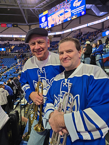 An man in a hat and a brown-haired man wear blue-and-white hockey jerseys and hold trumpets.