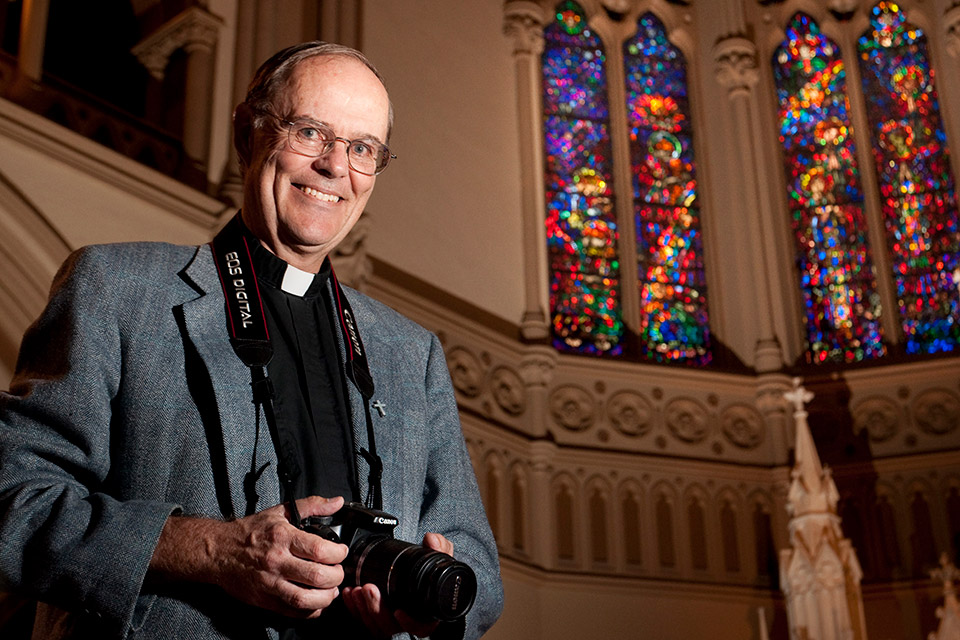 An older white man wearing a Roman collar and a camera around his neck stands near stained glass windows.