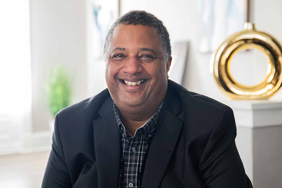 A smiling man in a suit jacket and checked shirt sits faces the camera with a living room in the background