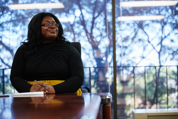 A graduate student holds a pen and paper at a table in a conference room in front of a large glass window while listening to a professor and smiling