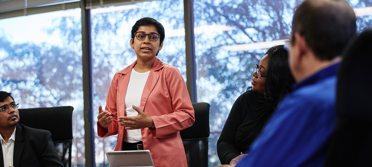 A female student stands before a group sitting around a table. She is talking to the group, who is listening attentively.