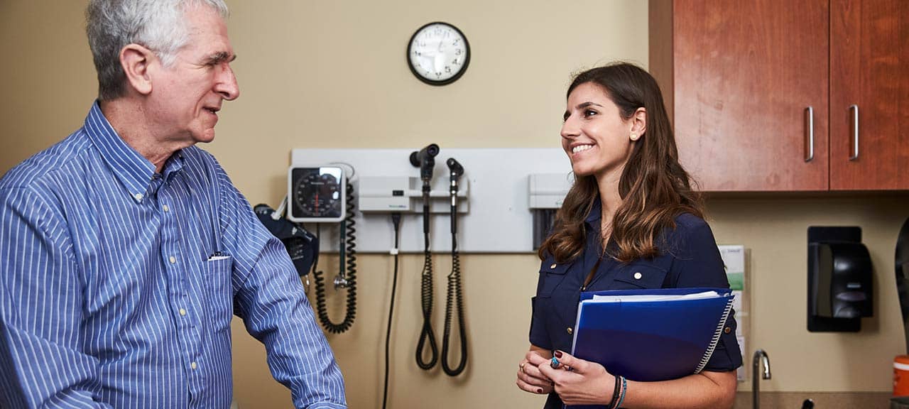 A female public health student speaks with an older male patient. The patient is sitting on the examination table in a doctor’s office while the student stands to speak with him while holding a notebook.