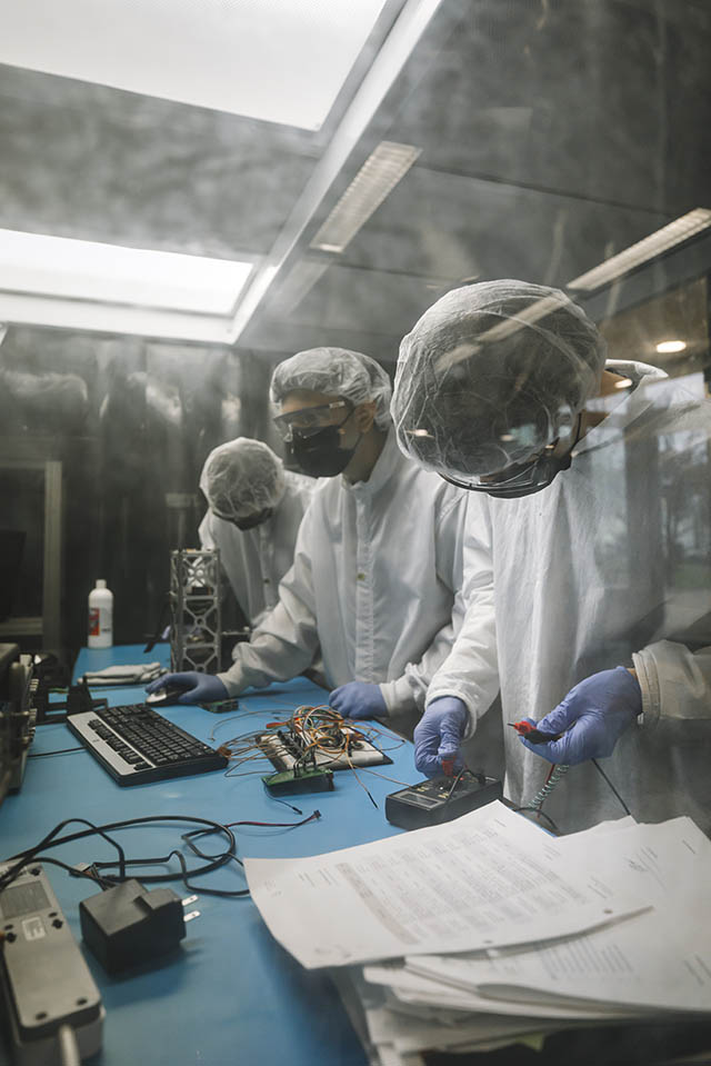 Three students wearing protective lab gear work at a table with various pieces of equipment and a computer.